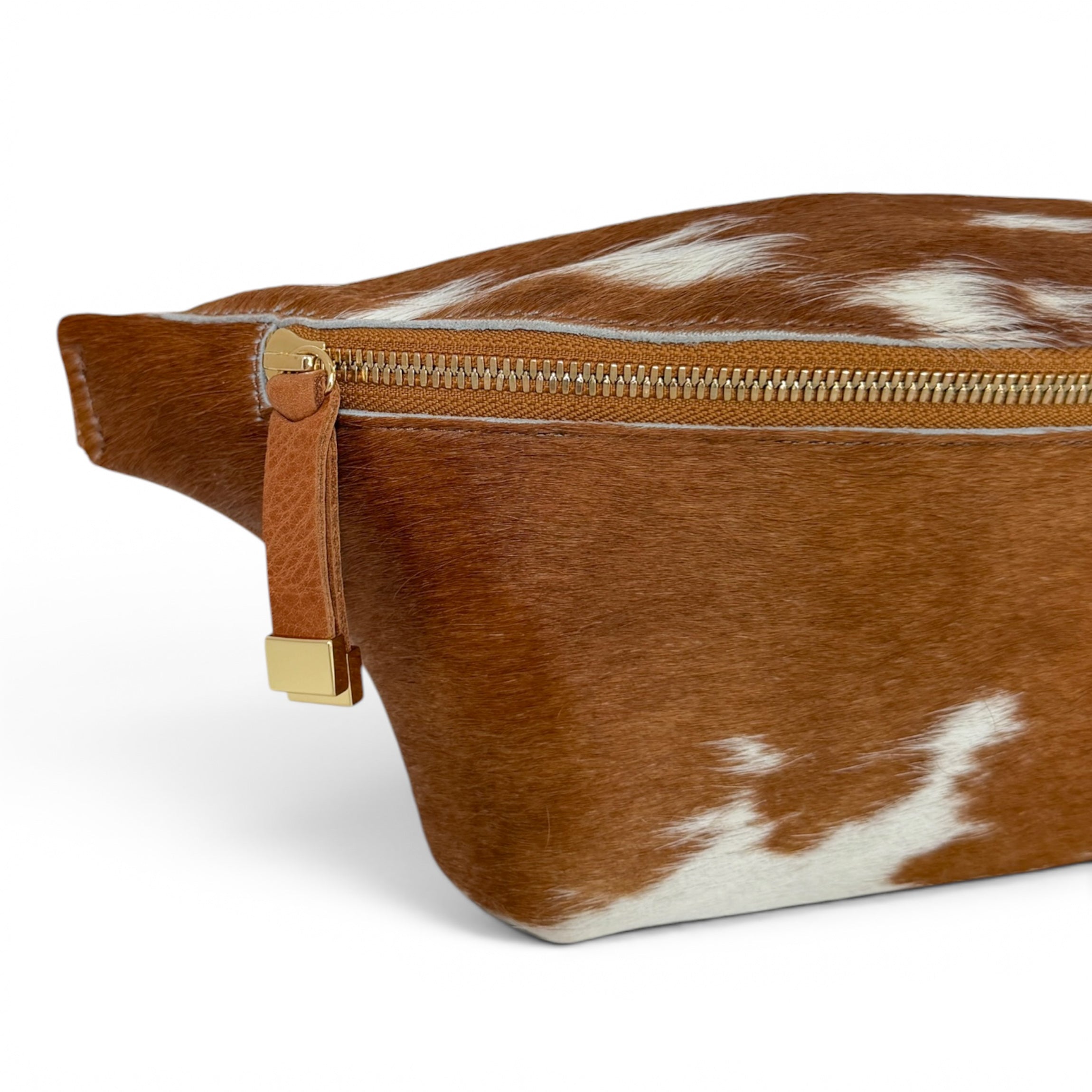 A close-up of a brown and white cowhide pouch with a gold-tone zipper and leather pull tab on a white background.